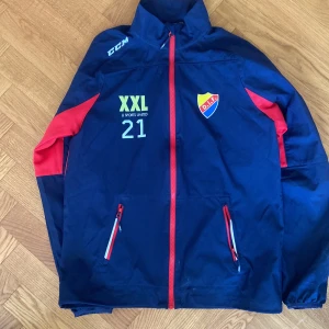 Blå vindjacka CCM DIF S - Sportig blå vindjacka från CCM med DIF-emblem på bröstet och röda detaljer längs dragkedja och ärmar. Jackan har tryck med XXL Sports United och siffran 21 på framsidan. Utrustad med två fickor med dragkedja och hög krage. Perfekt för dig som gillar hockey och Djurgården.