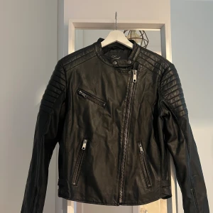 Svart skinnjacka från Xoul Edition - Snygg svart skinnjacka från Zoul Edition med coola quiltade detaljer på axlarna och flera dragkedjor framtill. Jackan har ståkrage med tryckknappar och en asymmetrisk dragkedja. Liten rivskada på ryggen, visas på bild. 