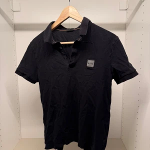 Svart slim fit skjorta från BOSS - Svart kortärmad skjorta från BOSS med slim fit-passform. Skjortan har klassisk krage, knappar framtill och en snygg BOSS-logga på bröstet. Tillverkad i mjuk bomull som känns skön mot huden. Perfekt för dig som gillar stilrena och enkla plagg.