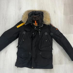 Svart Parajumpers dunjacka med päls - Svart Parajumpers dunjacka från Masterpiece Series med fluffig päls på huvan, coola patchar och orange detaljer. Jackan har flera fickor, dragkedja och knappar, quiltad insida och långärmad design. Perfekt för dig som vill ha en varm och snygg vinterjacka.