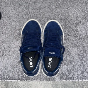 Dior b33 - Snygga blå sneakers från Dior med ovandel i mocka och canvas. Sidorna har det klassiska Dior-mönstret i grått och vitt, och skorna har vita sulor med unikt mönster undertill. Tjocka blå skosnören och rund tå ger en clean och lyxig vibe.(42 fast passar även 43)