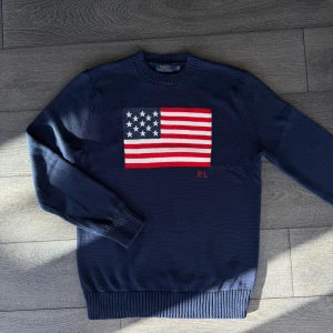 Mörkblå stickad tröja Polo Ralph Lauren - Mörkblå stickad tröja från Polo Ralph Lauren med stor amerikansk flagga på bröstet och RL-broderi. Klassisk rund halsringning och ribbade muddar. Perfekt för dig som vill sticka ut med en ikonisk look.