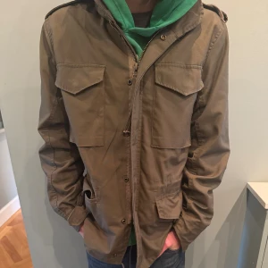 Olivgrön fieldjacka med avtagbart foder - Säljer en olivgrön parkajacka från Brandit med fyra stora fickor framtill, axelklaffar och huva. Jackan har dragkedja och knappar samt quiltat innerfoder som går att ta ur. Perfekt för lager-på-lager. 
