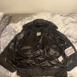 Svart pufferjacka från Moncler - Säljer en svart pufferjacka från Moncler med glansig finish och stor huva där Moncler-loggan är tryckt i vitt. Jackan har dragkedja och coolt serietidningsmärke på insidan. Perfekt för dig som vill ha en varm och snygg vinterjacka med streetkänsla.