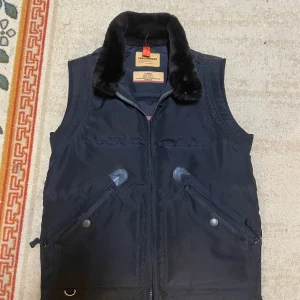 Svart dunväst från Parajumpers XS - Svart dunväst från Parajumpers i slim fit med mjuk pälsimitation på kragen. Västen har två snedställda fickor med dragkedja och detaljer i skinn, samt en snygg patch på nacken. Perfekt för kalla dagar och har en quiltad design. Storlek xs passar S