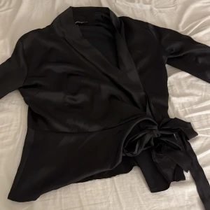 Svart omlottblus i satin - Säljer en svart omlottblus från NA-KD i glansigt satinmaterial. Blusen har långa ärmar med knapp vid ärmslut och en bred knytning i midjan som ger en snygg siluett. Perfekt för dig som gillar stilrena och trendiga plagg med lite extra lyxig känsla.