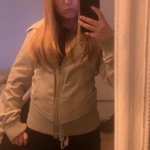 Beige jackamed från vila - Säljer en beige jacka med bred krage, dragkedja framtill och ribbade muddar vid ärmslut och nederkant. Jackan har snörning nedtill och en snygg passform. Använd fåtal gånher, inga hål eller missfärgningar. Jackan är i storlek M men passat dig som har XS eller S