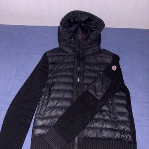 Moncler Cardigan - Svart pufferjacka från Moncler med stickade ärmar och huva. Jackan har dragkedja framtill, ribbade muddar och Moncler-logga på ena ärmen. Kombinerar glansigt puffer-material med grovstickade detaljer för en riktigt clean look.