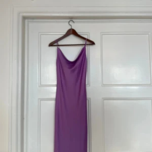 Lila långklänning med smala axelband - Säljer en lila långklänning med smala axelband och snygg draperad urringning. Klänningen har en slank siluett och är tillverkad i ett glansigt, mjukt material som ger en cool och elegant vibe. Perfekt för dig som gillar minimalistisk stil och vill sticka ut med färg.