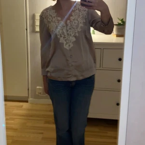 Beige blus med spets från H&M - Supersöt beige blus från H&M med v-ringning och vit spetsdetalj framtill. Blusen har långa ärmar, knappar framtill och är gjord i ett mjukt, lite glansigt material. 