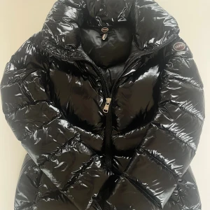 Svart Colmar pufferjacka med glans - Säljer en svart pufferjacka från Colmar med glansig finish och hög krage!💕 Jackan har Colmar-logga på ärmen och dragkedja framtill. Fylld med naturligt dun för extra värme och har en riktigt cool, puffig look som sticker ut!💕