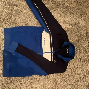 Blå sportig ziptröja från Lacoste - Sportig ziptröja från Lacoste i blått, vitt och mörkblått. Tröjan har dragkedja hela vägen, hög krage och klassisk Lacoste-logga på bröstet. Snygga färgblock och broderad text på framsidan. Perfekt för en chill och aktiv stil.
