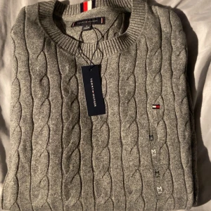 Grå kabelstickad tröja Tommy Hilfiger - Snygg grå kabelstickad tröja från Tommy Hilfiger med rund hals och diskret logga broderad på bröstet. Tröjan har ribbade muddar och är tillverkad i mjuk bomull, perfekt för att hålla värmen under kyliga dagar. Nypris 900kr, pris kan diskuteras!!
