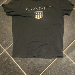 Svart GANT t-shirt med tryck storlek 152cm - Svart t-shirt från GANT med stort vitt logotryck och sköld med stjärnor och ränder på bröstet. Klassisk rund hals och korta ärmar. Tillverkad i mjuk bomull som är skön att ha på sig. Perfekt för en chill och stilren look. Storlek 152cm