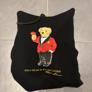Svart hoodie Polo Bear Ralph Lauren M - Svart hoodie från Polo Ralph Lauren med ikonisk Polo Bear-tryck på bröstet. Insidan av huvan har ett rött och gult rutigt mönster i mjuk fleece. Tröjan har snörning vid huvan och är långärmad. Perfekt för dig som gillar streetwear med lite extra attityd.