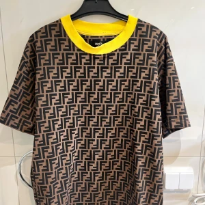 Fendi t-shirt med FF-mönster och gul krage - Snygg t-shirt från Fendi med ikoniskt FF-logomönster i svart på brun bakgrund och en fet gul ribbad krage. Oversized fit och korta ärmar. Tillverkad i mjuk bomull som känns skön mot huden. Perfekt för dig som vill sticka ut med en lyxig streetstyle.