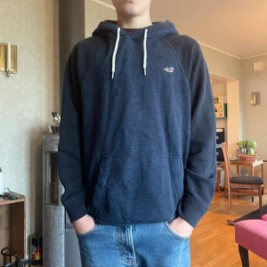 Mörkblå hoodie från Hollister - Säljer en mörkblå hoodie från Hollister med vit snörning och klassisk logga på bröstet. Tröjan har känguruficka framtill och långa ärmar. Perfekt för dig som gillar en avslappnad och stilren look.