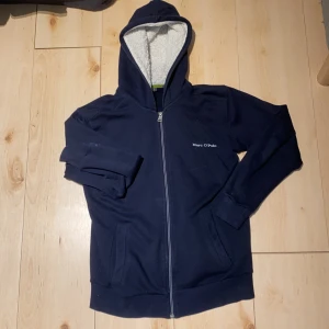 Mörkblå hoodie från Marc O'Polo - Mörkblå hoodie från Marc O'Polo med vit text på ryggen och liten logga fram. Dragkedja hela vägen, två fickor och mysigt vitt teddyfoder i huvan. Perfekt för kyliga dagar och har en klassisk, avslappnad look. Hål på armbågen, går enkelt att laga om man kan sy.