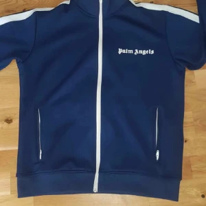 Blå track jacket från Palm Angels - Snygg marinblå track jacket från Palm Angels med vita detaljer och dragkedja framtill. Jackan har vita ränder längs axlarna, två fickor med dragkedja och logga på bröstet. Tillverkad i 100% polyester för en sportig vibe.