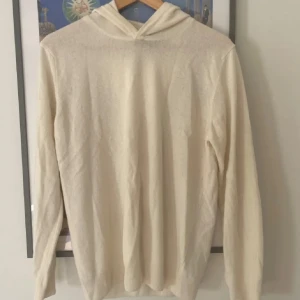 Beige hoodie i tunn stickad ull - Säljer en stilren beige hoodie i tunn stickad ull. Tröjan har lång ärm, huva och en avslappnad passform. Perfekt för lager-på-lager under hösten eller vintern. Enkel och clean look som passar till det mesta.