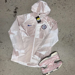 Chelsea x Nike vindjacka med mönster - Snygg vindjacka från Nike i samarbete med Chelsea FC. Jackan har ett unikt abstrakt mönster i beige och vitt, hel dragkedja framtill, huva och Chelsea-logga på bröstet. Perfekt för dig som vill sticka ut med sportig stil.