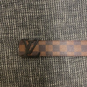 Louis Vuitton rutigt brunt bälte - Säljer ett Louis Vuitton bälte med klassiskt LV-spänne i svart metall. Bältet har ett rutigt mönster i bruna och mörkbruna nyanser och är gjort i skinn. Perfekt accessoar för att lyfta din outfit med en lyxig touch.