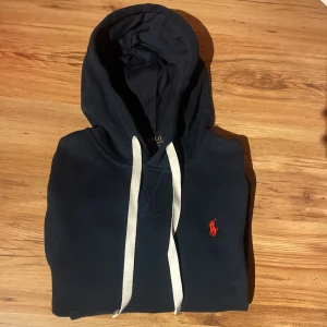 Mörkblå hoodie från Polo Ralph Lauren - Säljer en mörkblå hoodie från Polo Ralph Lauren med vit snörning och klassisk röd logga broderad på bröstet. Tröjan har huva, känguruficka och ribbade muddar. Perfekt för dig som gillar stilren streetwear med premiumkänsla.
