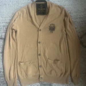 Beige kofta från Morris  - Stilren beige kofta från Morris med broderad logga på bröstet och knäppning framtill. Tröjan har två fickor och ribbade muddar vid ärmslut och nederkant. Perfekt för lager-på-lager och snygg till jeans eller chinos.