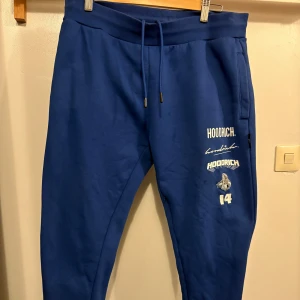 Blå Hoodrich mjukisbyxor  - Blå mjukisbyxor från Hoodrich med vita tryckdetaljer och broderad logga bak. Byxorna har snörning i midjan, sidofickor och en bakficka. Skön passform och streetwear-vibe, perfekta för chill eller sportig stil. Large men passar medium 