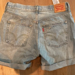 Levi's ljusblå jeansshorts W30 - Säljer ett par ljusblå jeansshorts från Levi's med klassisk femficksdesign, uppvikta ben och slitna detaljer framtill. Shortsens midja är normal och de har knappar i gylfen. Perfekt för dig som gillar en chill och avslappnad stil.