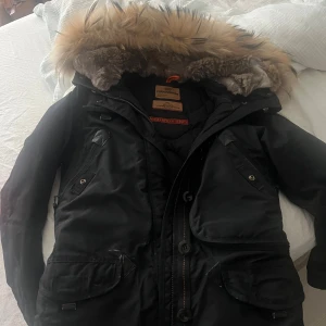 Svart Parajumpers vinterjacka med päls - Svart Parajumpers vinterjacka med stor huva och päls, ikonisk gul rem på axeln och patch på ärmen. Jackan har flera fickor, dragkedja och knappar framtill. Slim fit-modell, perfekt för kalla dagar.