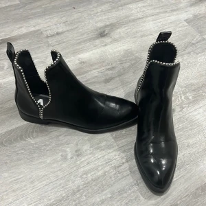 Svarta chelsea boots med pärldetaljer Zara  - Snygga svarta chelsea boots i skinn med rund tå och platt sula. Runt öppningen sitter coola silverfärgade pärldetaljer som ger extra edge. Bootsens klassiska form gör dem lätta att matcha och de har dragflik baktill för enkel påtagning.