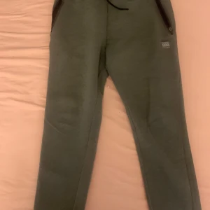 Gröna mjukisbyxor från Superdry Sport - Snygga gröna mjukisbyxor från Superdry Sport i storlek medium. Byxorna har dragkedjeförsedda fickor, snörning i midjan och en liten logga på framsidan. Materialet är en mix av bomull, polyester och elastan för skön stretch och passform.