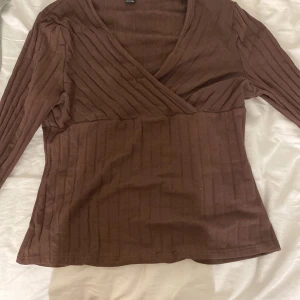 Brun omlottop med ribbad struktur - Säljer en brun långärmad omlottop med ribbad struktur. Toppen har v-ringning och sitter snyggt med en figurnära passform. Perfekt för dig som gillar en enkel men stilren look. Hade gärna behållt den men är för liten för den 