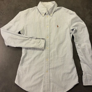 Randig skjorta från Polo Ralph Lauren XS - Klassisk feminin skjorta från Polo Ralph Lauren i vitt och blått med smala ränder. Custom fit-modell med lång ärm, knappar framtill och broderad logga på bröstet. 