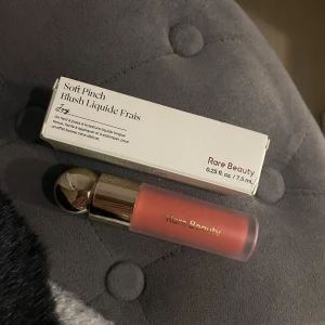 Rare Beauty Soft Pinch Liquid Blush Joy - Helt ny Rare Beauty Soft Pinch Liquid Blush i nyansen Joy, en flytande rouge i en varm persikoton. Kommer i en snygg frostad tub med guldig kork. Lätt att applicera och blenda för ett fräscht glow. Perfekt för dig som vill ha en naturlig och fräsch look.