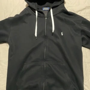 Svart hoodie från Polo Ralph Lauren (Limited edition) - Svart hoodie från Polo Ralph Lauren med vit broderad logga på bröstet. Tröjan har hel dragkedja, vita snören i huvan och känguruficka framtill. Klassisk och stilren design som passar perfekt till chill eller streetwear. Pris kan diskuteras, eftersom vill bara bli av med tröjan, bara och höra av dig!🍾🍾