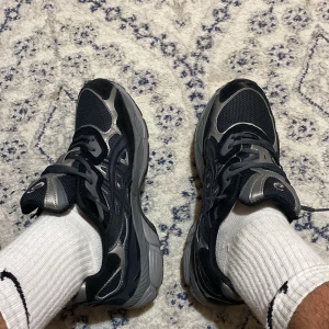 Svarta och silver Asics sneakers - Säljer ett par snygga Asics sneakers i svart och silver. Skorna har mesh- och syntetpaneler, klassisk snörning och rund tå. Perfekta för dig som gillar sportig stil och vill ha bekväma skor till vardagen.