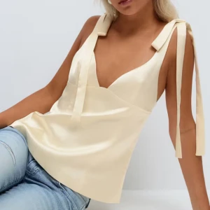 Söt topp - Supersöt ljusgul topp från Gina Tricot i glansig satin. Toppen har breda axelband med knyt, v-ringning och figurnära passform. Perfekt att styla med jeans eller kjol för en trendig look. Dragkedja i ryggen för enkel påklädning. Smink fläck på ryggen (se bild) går säkert att ta bort! 