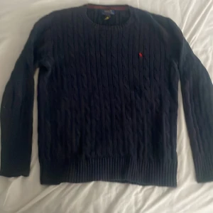 Mörkblå kabelstickad tröja Polo Ralph Lauren - Kabelstickad mörkblå tröja från Polo Ralph Lauren med klassisk rund hals och röd broderad logga på bröstet. Tröjan har långa ärmar och ribbade muddar vid ärmslut och nederkant. Perfekt för dig som gillar preppy och stilren look. Är öppen för frågor och priset är ej hugget i sten 