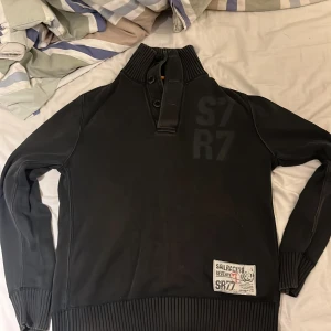 Svart Sail Racing tröja med hög krage - Svart långärmad tröja från Sail Racing med hög ribbad krage och knappslå. Snygg detalj med patch och tryck på framsidan. Tröjan har breda ribbade muddar vid ärmslut och nederkant. Perfekt för dig som gillar stilren look. Denna blir väldigt snygg med ett par jeans och vikt krage den sitter väldigt bra. Pris går att diskutera😀