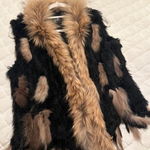 Svart och brun pälsväst från Hollies - Säljer en fluffig pälsväst från Hollies i svart med bruna och beige inslag. Västen har en bred krage i ljusbrun päls och coola detaljer med olika färgade pälsfläckar. Perfekt för att ge din outfit extra edge och värme under kyliga dagar.