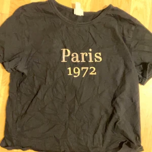 Svart t-shirt Paris 1972 H&M - Svart t-shirt från H&M med tryck i vitt och beige text 'Paris 1972' på bröstet. Klassisk rund halsringning och korta ärmar. Tillverkad i mjuk bomull, perfekt för en chill och stilren look.