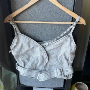 Grå denim korsett från Miss Sixty - Säljer en grå denim korsett från Miss Sixty med justerbara axelband och snygg omlott-design framtill. Toppen har ett brett bälte i midjan med spänne som ger en cool siluett. Perfekt för dig som gillar statement-plagg och streetstyle.