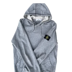 Grå hoodie från Stone Island - Snygg grå hoodie från Stone Island med klassisk logga på ärmen. Tröjan har huva med snörning och känguruficka framtill. Tillverkad i mjuk bomull för maximal komfort. Perfekt för dig som gillar streetwear och vill ha en stilren look.