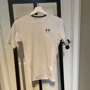 Vit kompressionst-shirt från Under Armour - Säljer en vit kompressionst-shirt från Under Armour med svart logga på bröstet. T-shirten är kortärmad och tillverkad i ett syntetmaterial som andas, perfekt för träning. Snygg och sportig passform som sitter tight mot kroppen.