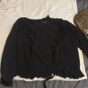 Svart bolerotopp från Stockh LM - Supersnygg svart bolerotopp från Stockh LM med omlottknytning och volangdetaljer längs kanterna. Stickad struktur med långa ärmar och breda muddar med knapp och band. Croppad passform som ger en trendig look till jeans eller kjol.