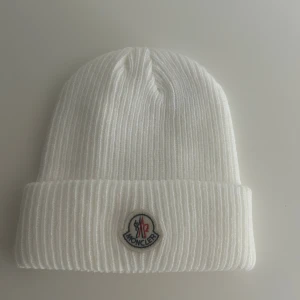 Vit ribbad mössa från Moncler - Säljer en stilren vit ribbstickad mössa från Moncler med klassisk uppvikt kant och broderad logotyp framtill. Mössan är tillverkad i mjukt material och passar perfekt för kyliga dagar. Inte alls använd för den var för liten.