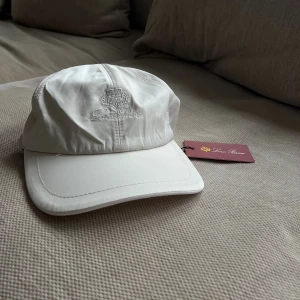 Vit keps från Loro Piana - Exklusiv vit keps från Loro Piana med broderad logga framtill. Kepsen har en klassisk böjd skärm och insidan är fodrad med rutigt tyg och orange kant. Tillverkad i ett slätt, lyxigt material och har elastisk baksida för skön passform.