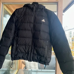 Svart pufferjacka från Adidas - Svart pufferjacka från Adidas med huva och dragkedja framtill. Jackan har två sidofickor med dragkedja och Adidas-logga på bröstet. Den är quiltad och har snörning nedtill för att justera passformen. Perfekt för kalla dagar.